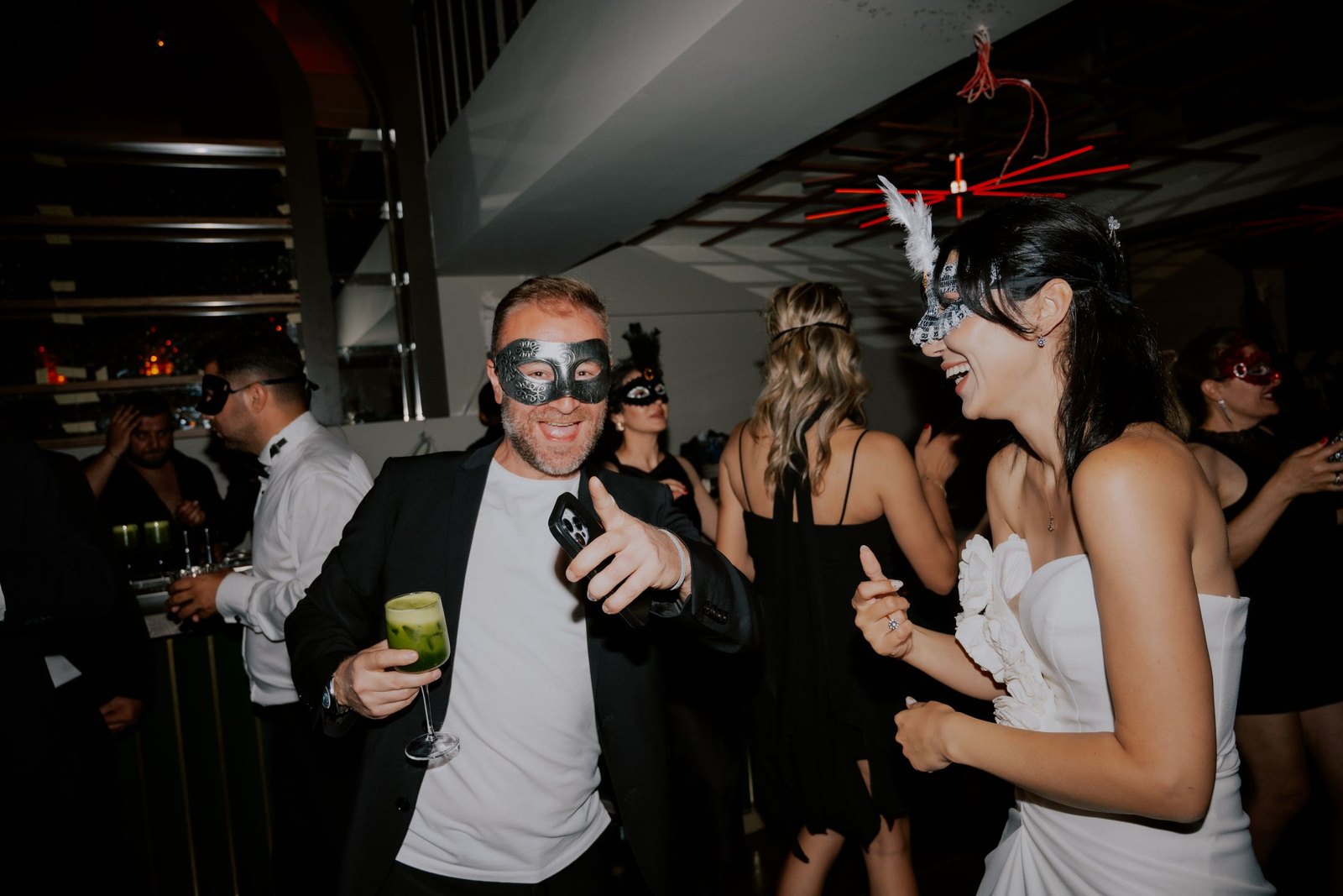 Guests in suits and evening dresses wearing masquerade masks, dancing with drinks at a nighttime party